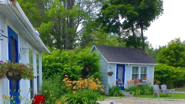 A cottage with a blue door, surrounded by a garden with various flowers and a wooden deck. - Isleview Motel and Cottages (Trenton)