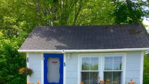 A small, single-story house with a blue door and white exterior, number 5, surrounded by a garden with various flowers and two wooden chairs outside. - Isleview Motel and Cottages (Trenton)