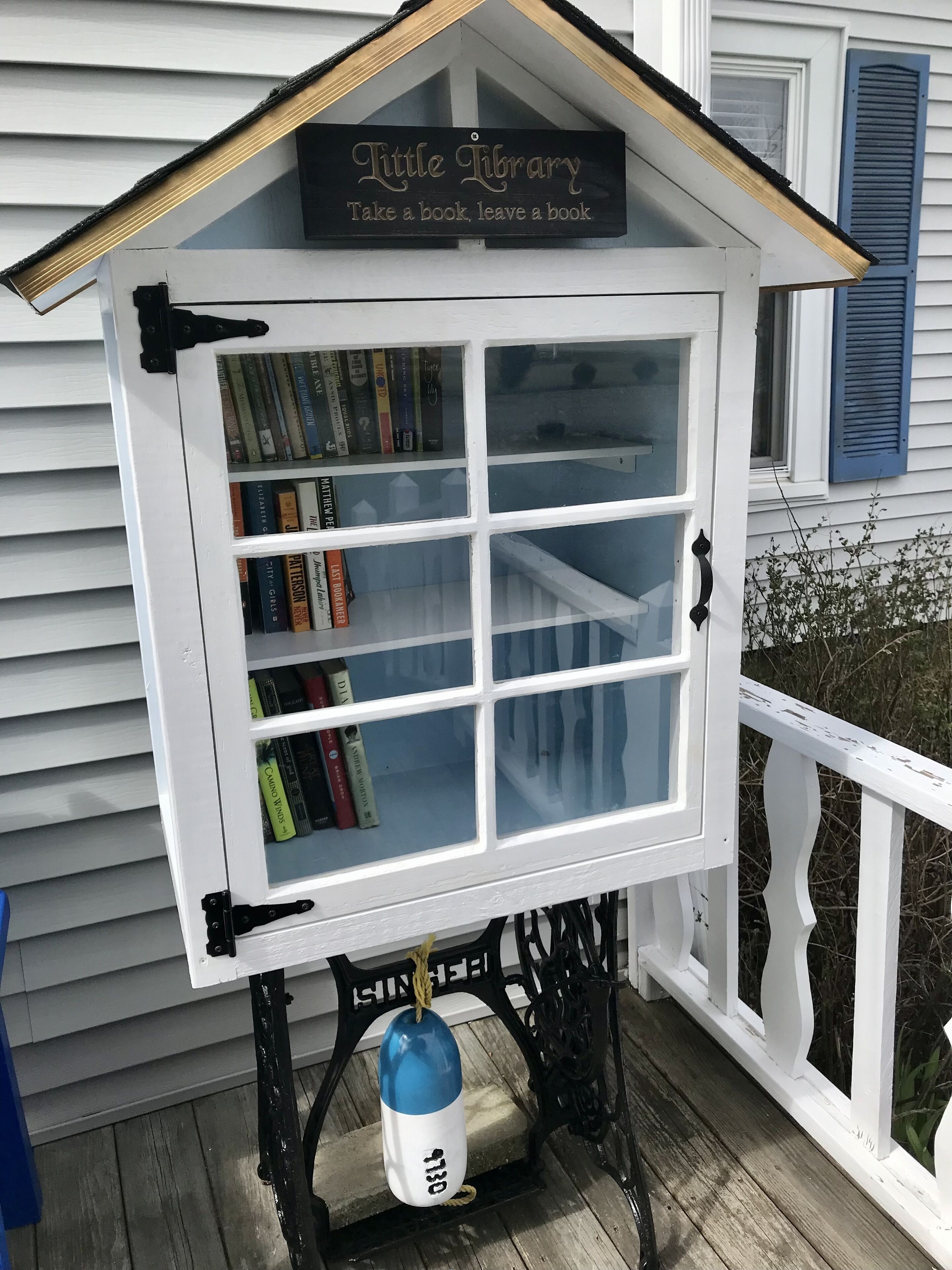 A small, white bookcase with a sign that reads "Little Library: Take a book, leave a book." It is mounted on a black metal stand with a rope tied around it.