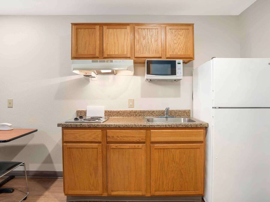A small kitchen with wooden cabinets, a microwave, a refrigerator, a sink, and a table with a chair.