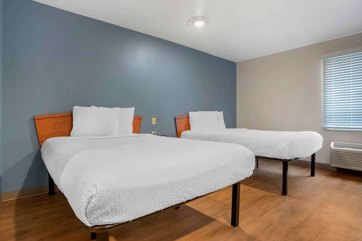 A hotel room with two beds, wooden flooring, and a window with blinds.