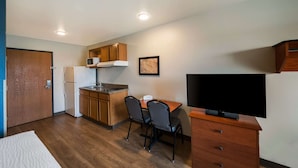 A compact kitchen with a microwave, refrigerator, and wooden cabinets. There is a dining table with two chairs and a flat-screen TV on a wooden dresser. - WoodSpring Suites Fort Worth Fossil Creek (Fort Worth)