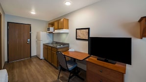 A compact kitchen with wooden cabinets, a refrigerator, a microwave, and a small dining table with a chair. - WoodSpring Suites Fort Worth Fossil Creek (Fort Worth)