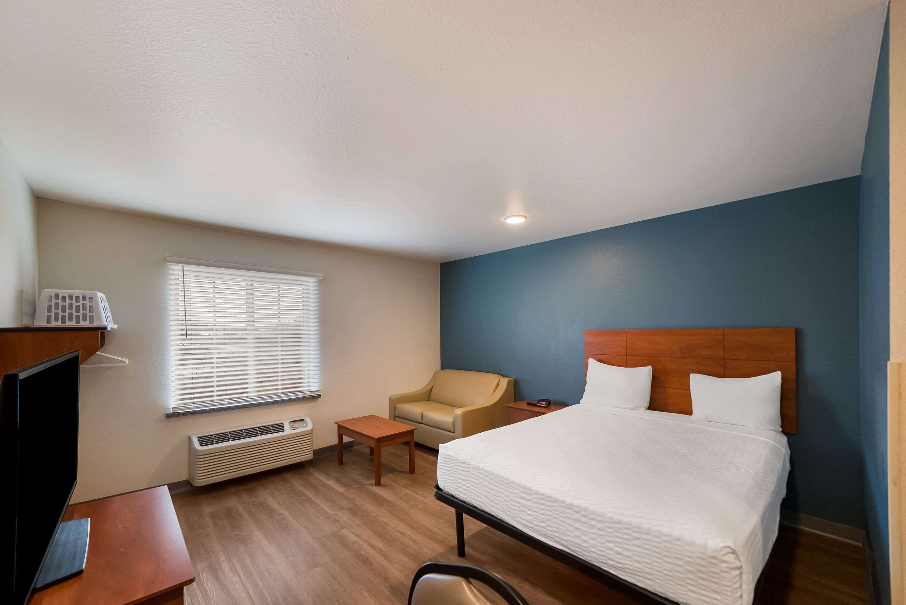 A hotel room with a bed, a desk, a chair, a TV, a heater, and a window with blinds.