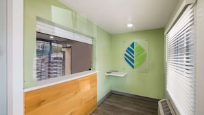 A reception area with a wooden counter, a glass partition, a green wall, and a vent. - WoodSpring Suites Fort Worth Fossil Creek (Fort Worth)