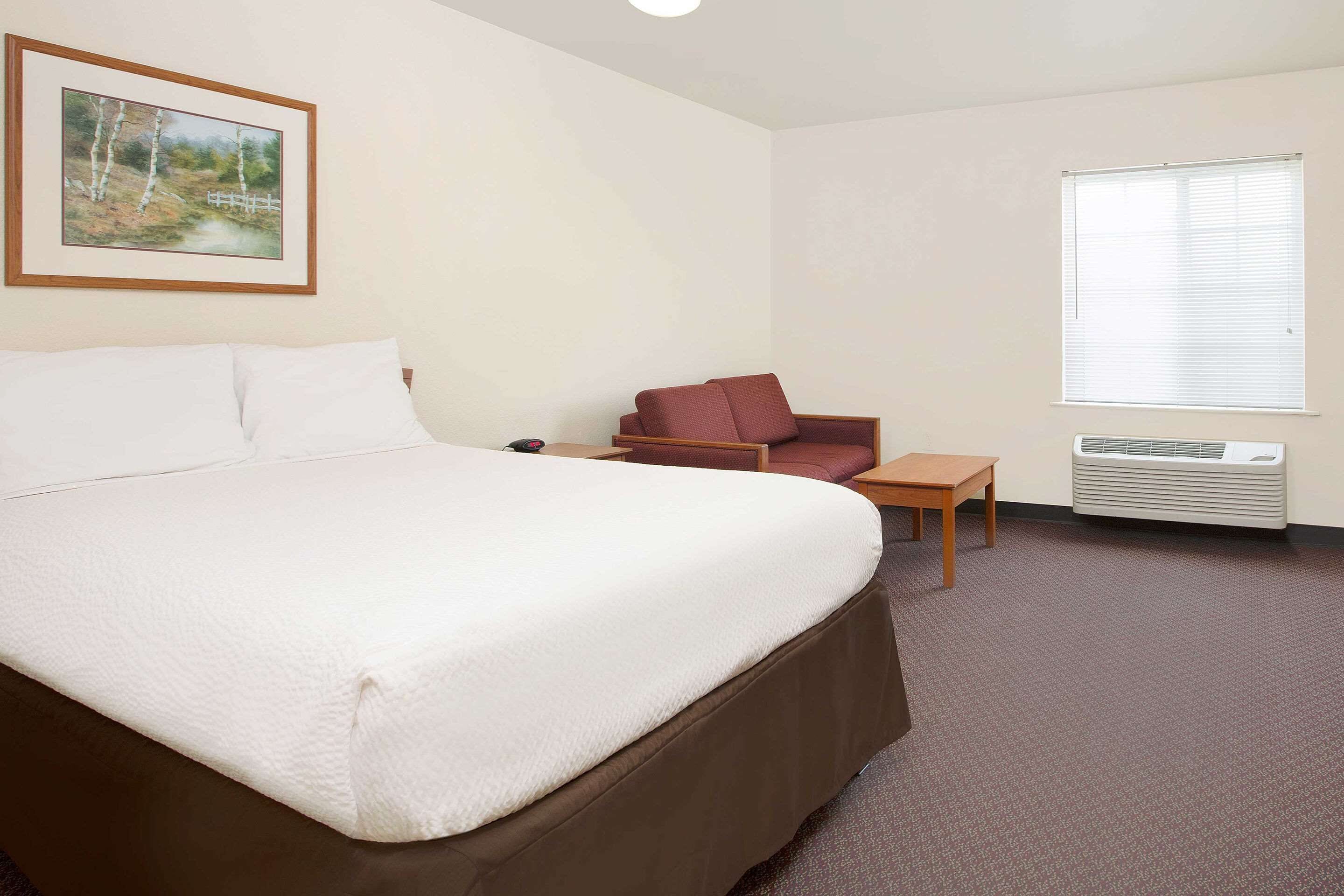 Een hotelkamer met een groot bed, een kleintje tafeltje, een rode stoel, een raam met jaloezieën en een lijst met een afbeelding aan de muur.