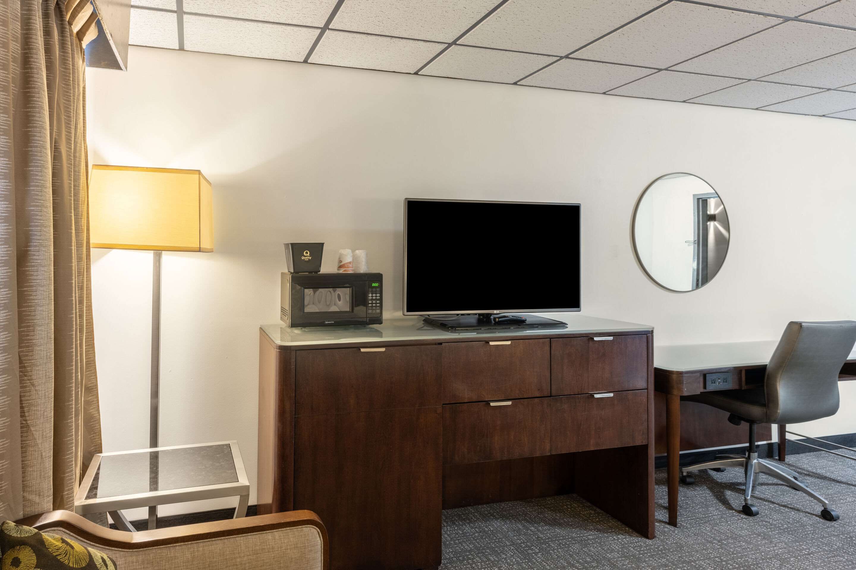 Ett hotellrum med ett skrivbord i trä, en platt-TV, en mikrovågsugn, ett litet bord, en stol, en lampa och en spegel.