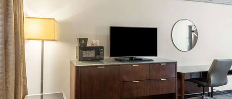 Ett hotellrum med ett skrivbord i trä, en platt-TV, en mikrovågsugn, ett litet bord, en stol, en lampa och en spegel.