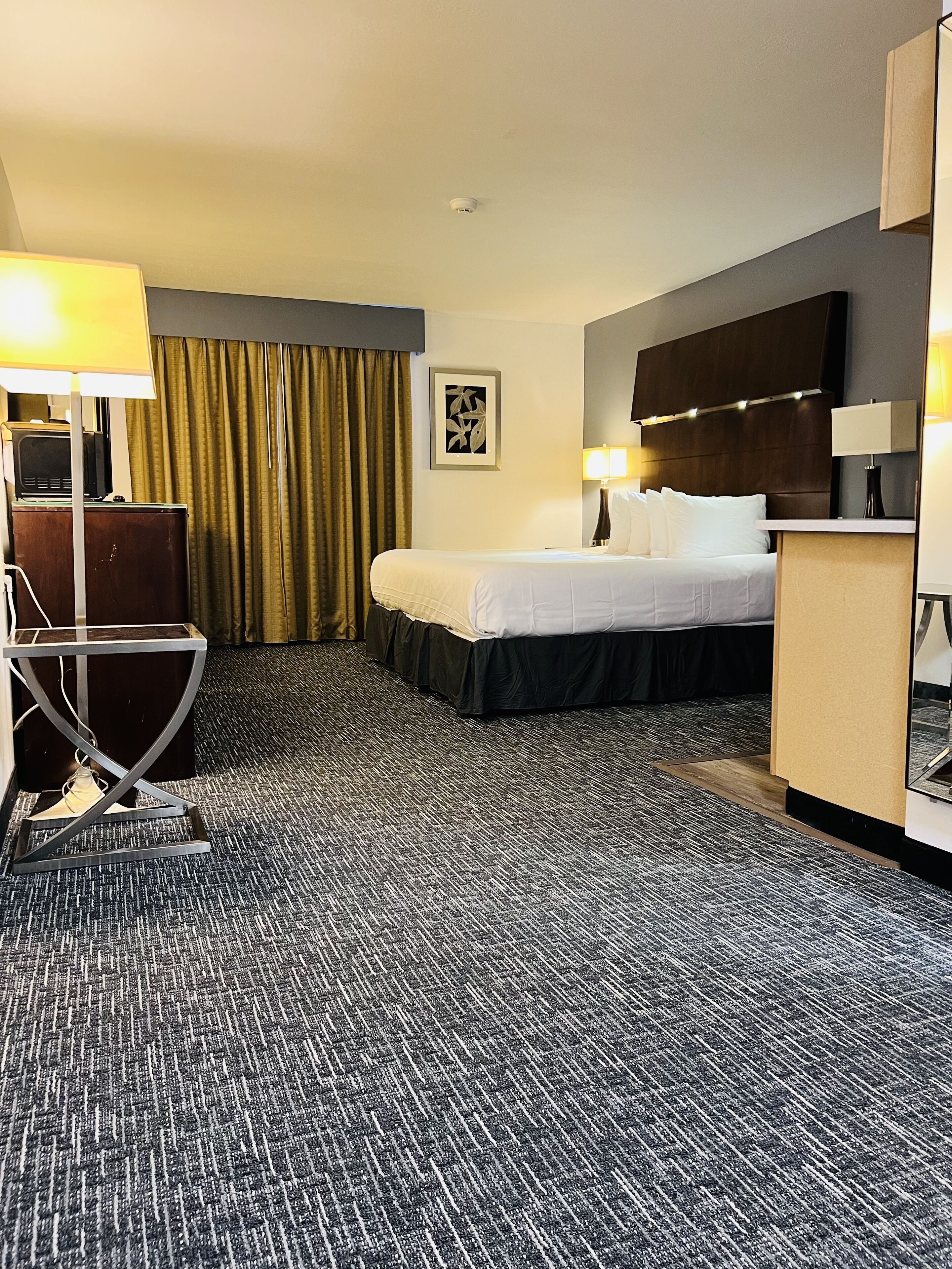 Une chambre d’hôtel avec un lit, un bureau, une chaise, une lampe, un tableau au mur et un luminaire au plafond.
