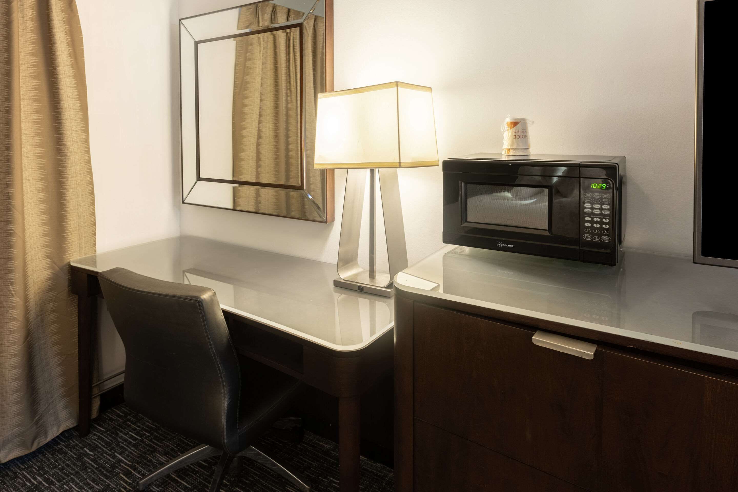 A hotel room with a desk, a chair, a lamp, a microwave, and a mirror.