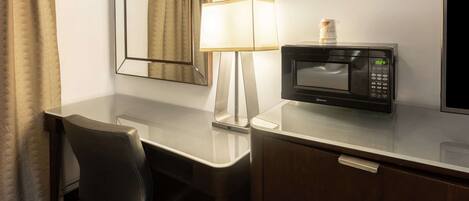 A hotel room with a desk, a chair, a lamp, a microwave, and a mirror.