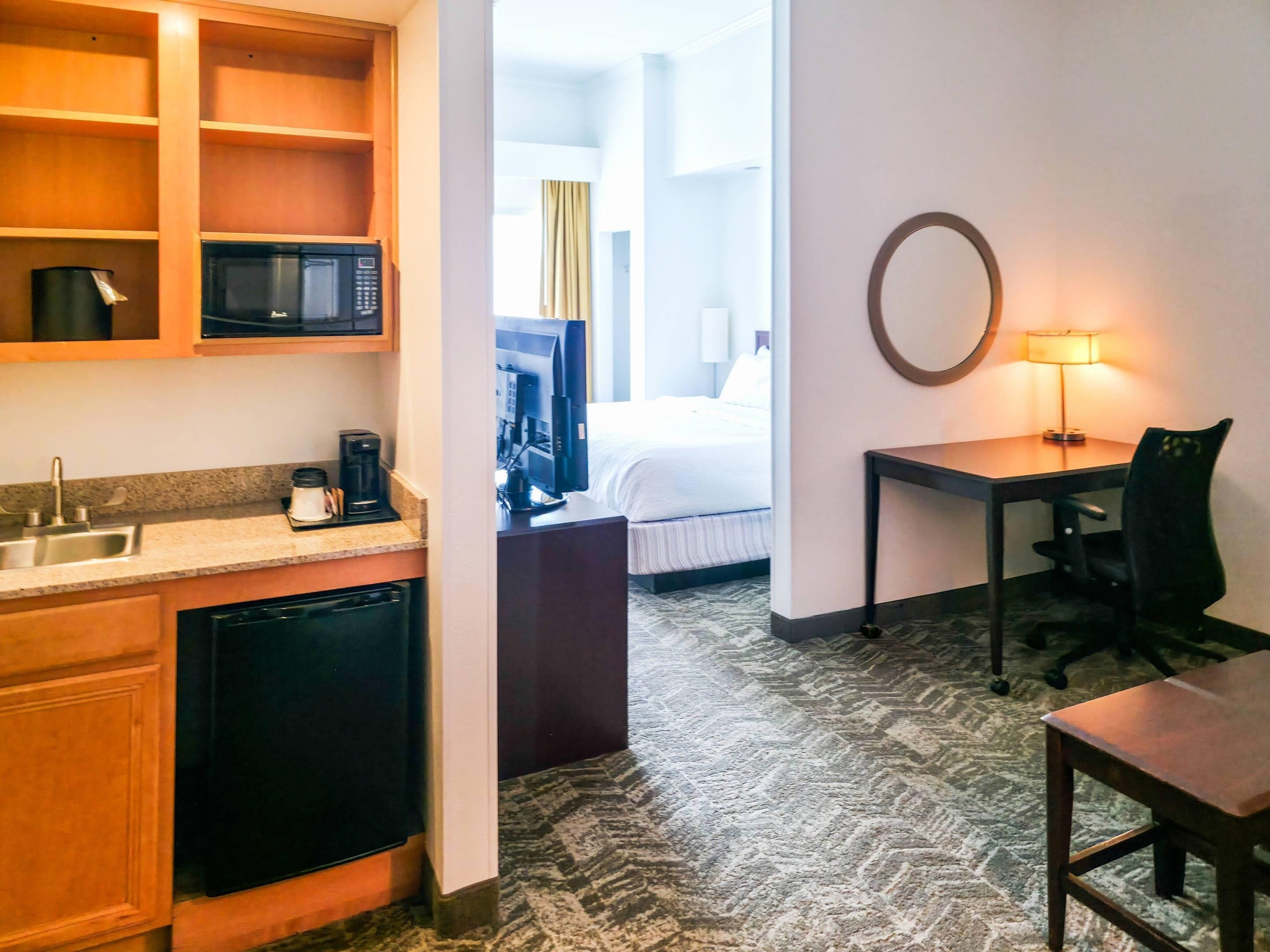 A hotel room with a kitchenette, a bed, a desk, and a lamp.