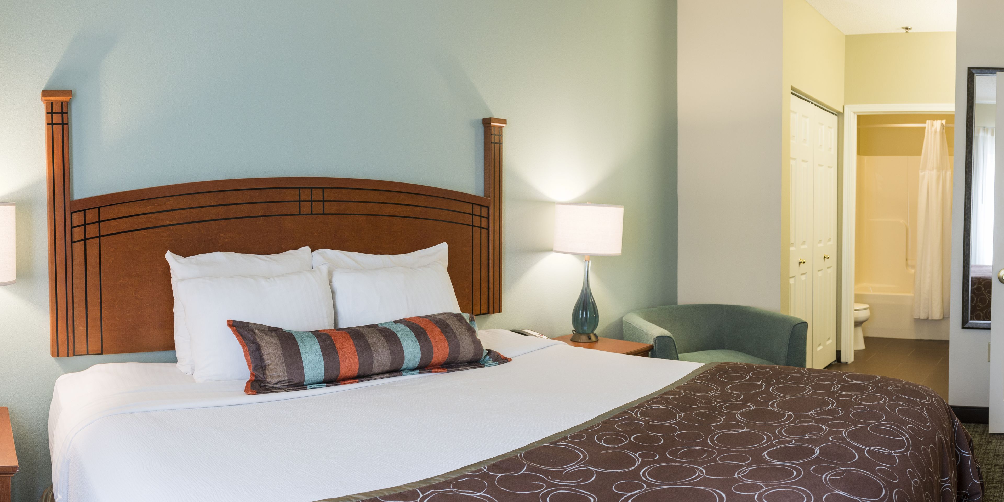 Habitación de hotel con una cama grande, una mesita de noche, una lámpara, una silla y un baño visible a través de una puerta abierta.