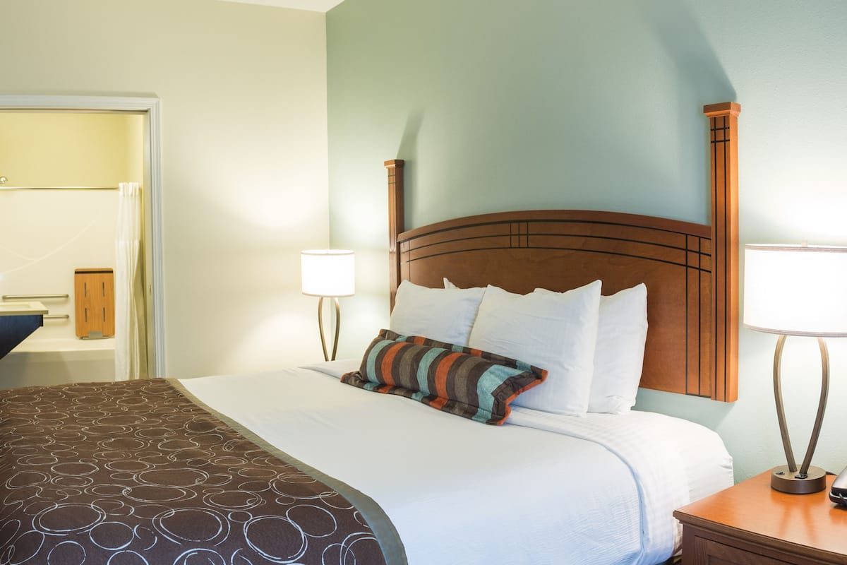 A hotel room with a bed, a nightstand, a lamp, and a door leading to a bathroom.