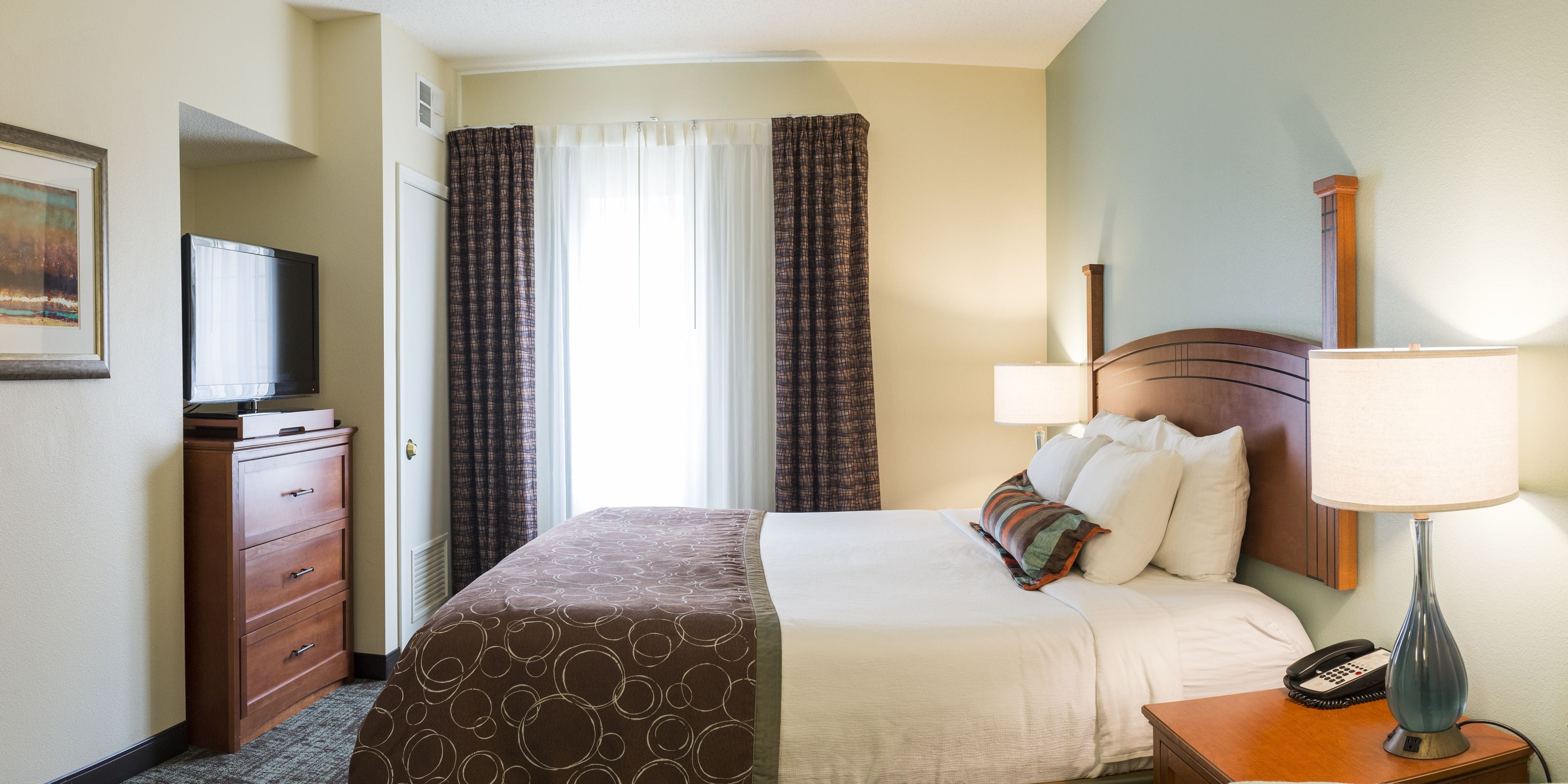 A hotel room with a bed, a dresser, a television, a lamp, and a window with curtains.