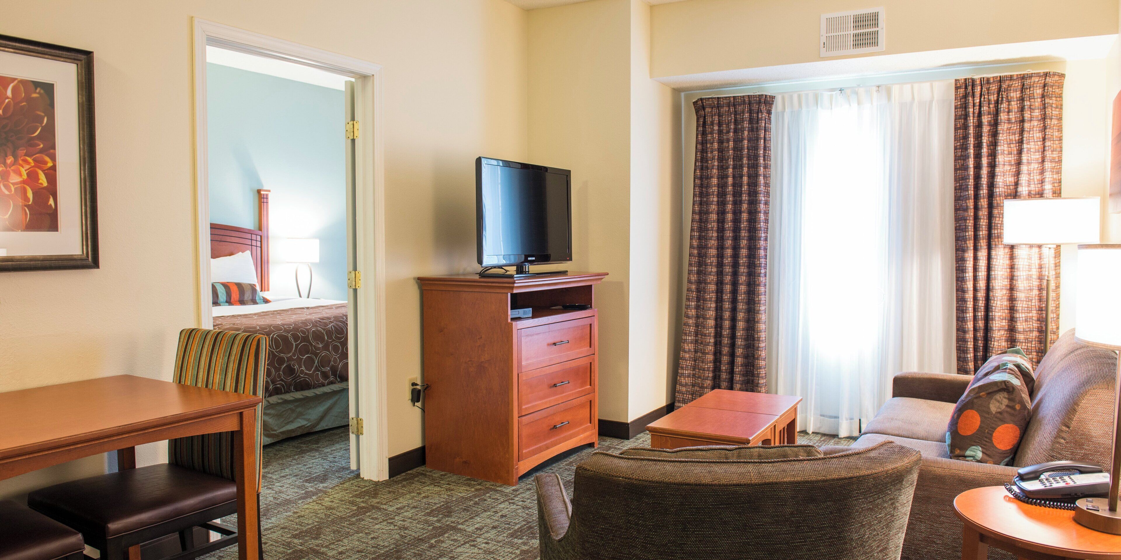 A hotel room with a television on a wooden stand, a desk with a chair, a sofa, a small table with a lamp, and a bed with a headboard.