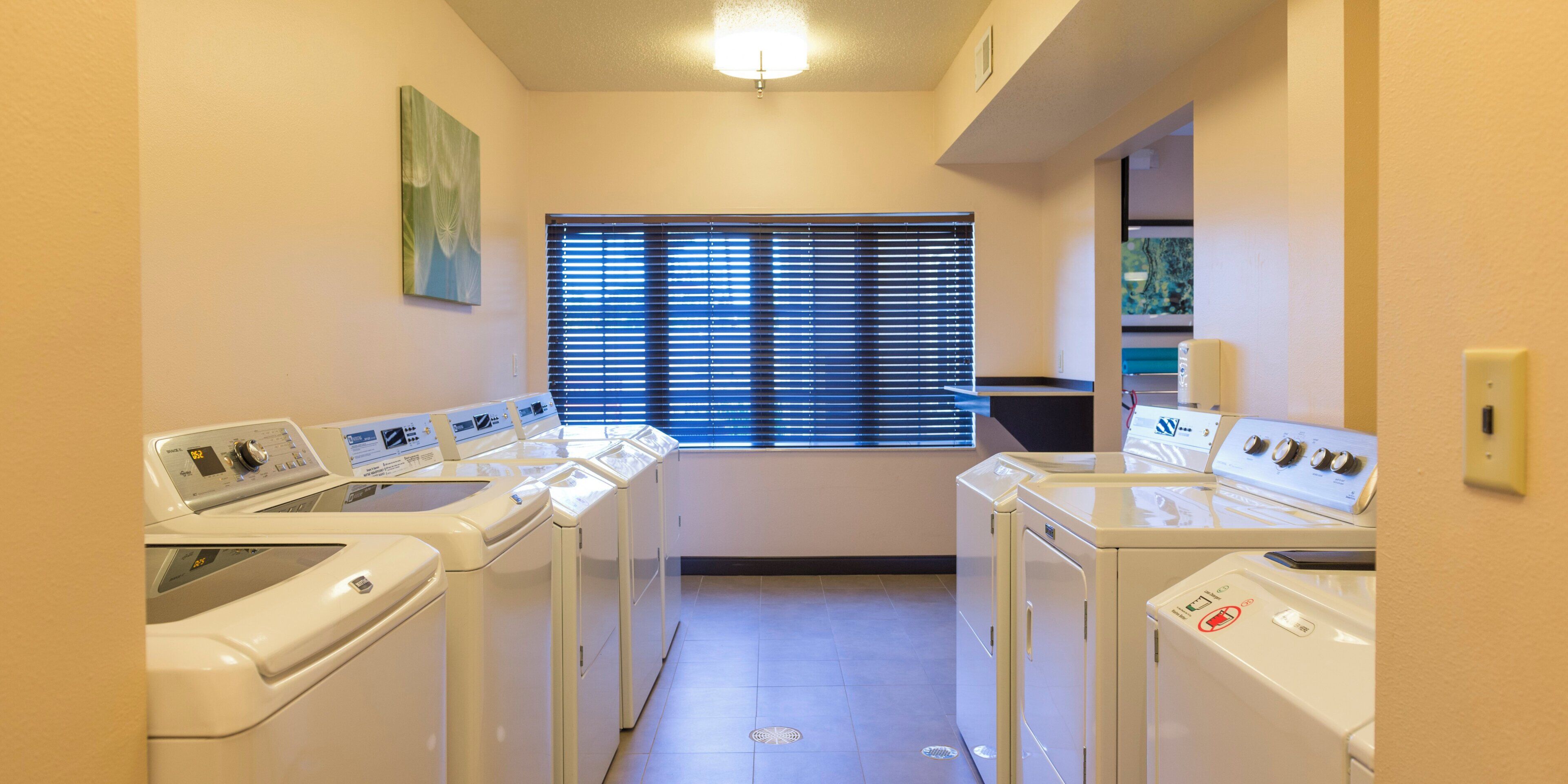 A laundry room with four front-loading washers and dryers, a window with blinds, and a painting on the wall.
