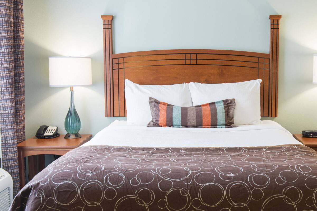 A hotel room with a wooden headboard, a large bed with a patterned bedspread, two bedside tables with lamps, a telephone, and a wall-mounted heater.