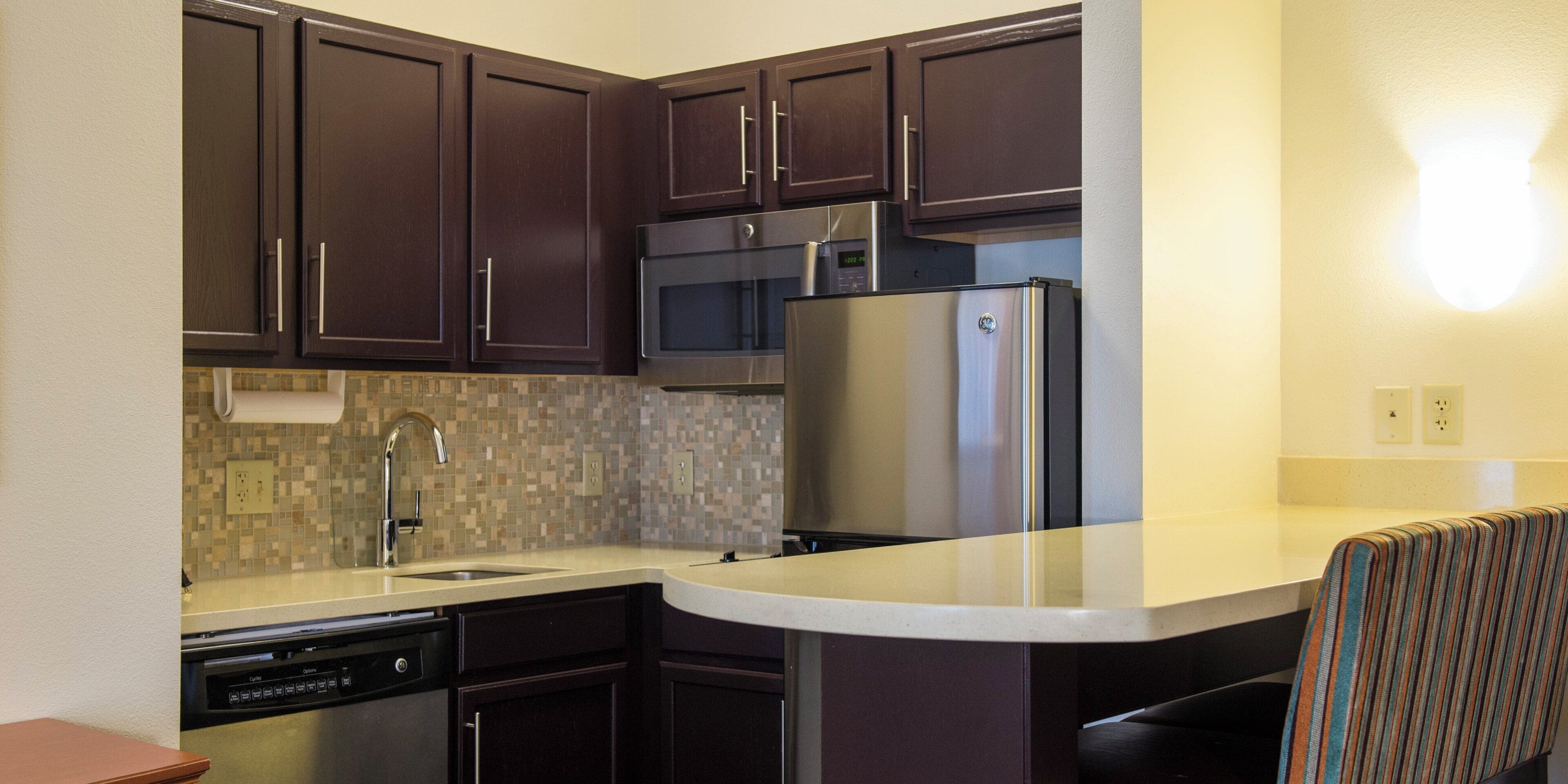 Una cocina compacta con gabinetes de madera oscura, un refrigerador de acero inoxidable, un microondas y un lavavajillas.