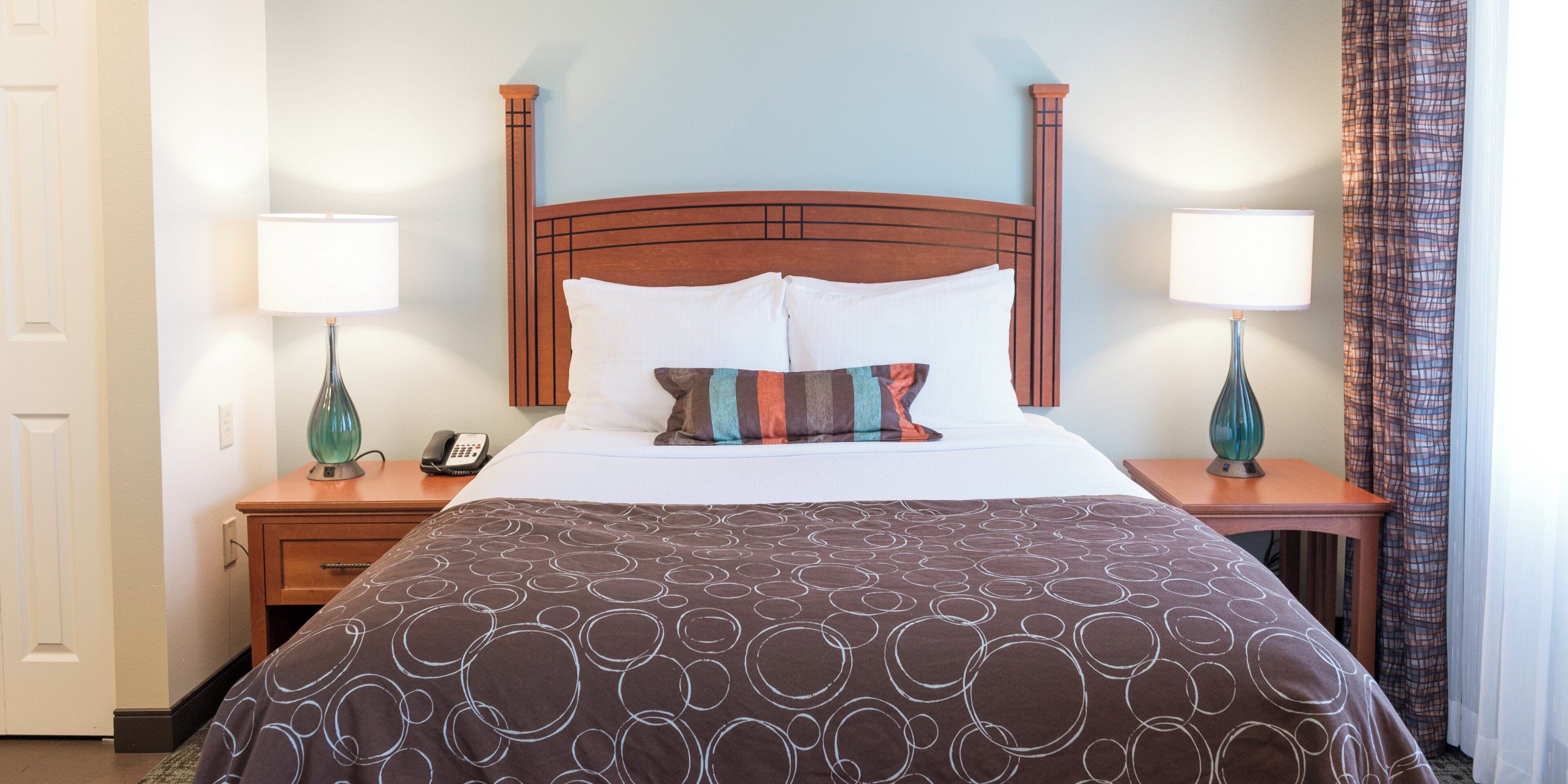 A hotel room with a wooden headboard, a bed with white pillows, a patterned throw, and two bedside lamps.