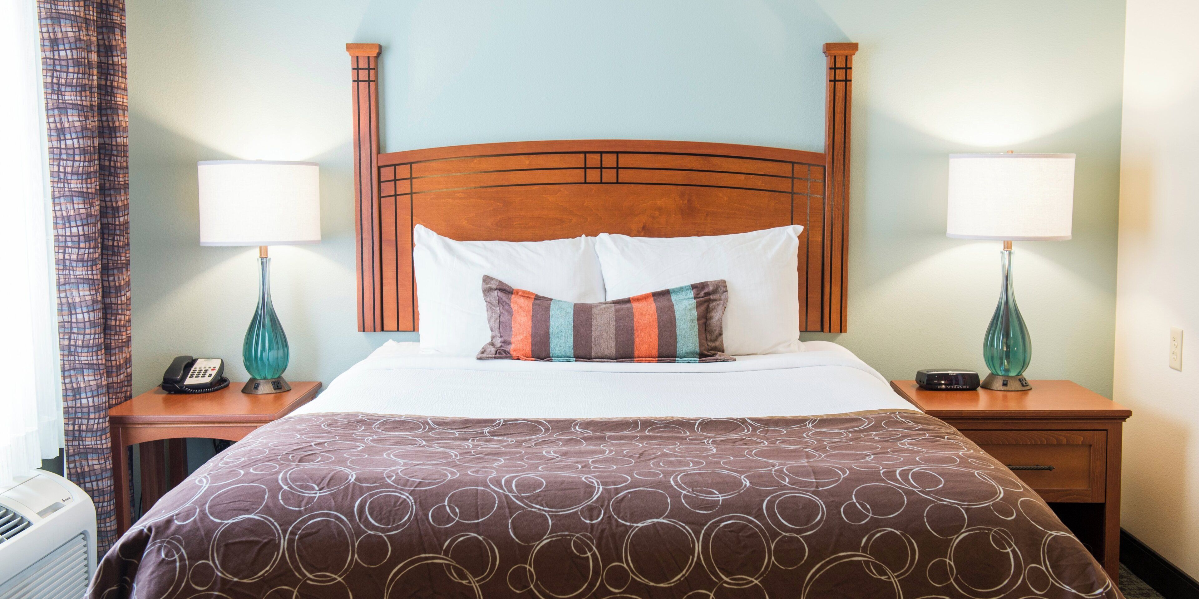 Habitación de hotel con cabecera de madera, cubrecama blanco, almohada a rayas, dos lámparas de noche y un teléfono en la mesita de noche.