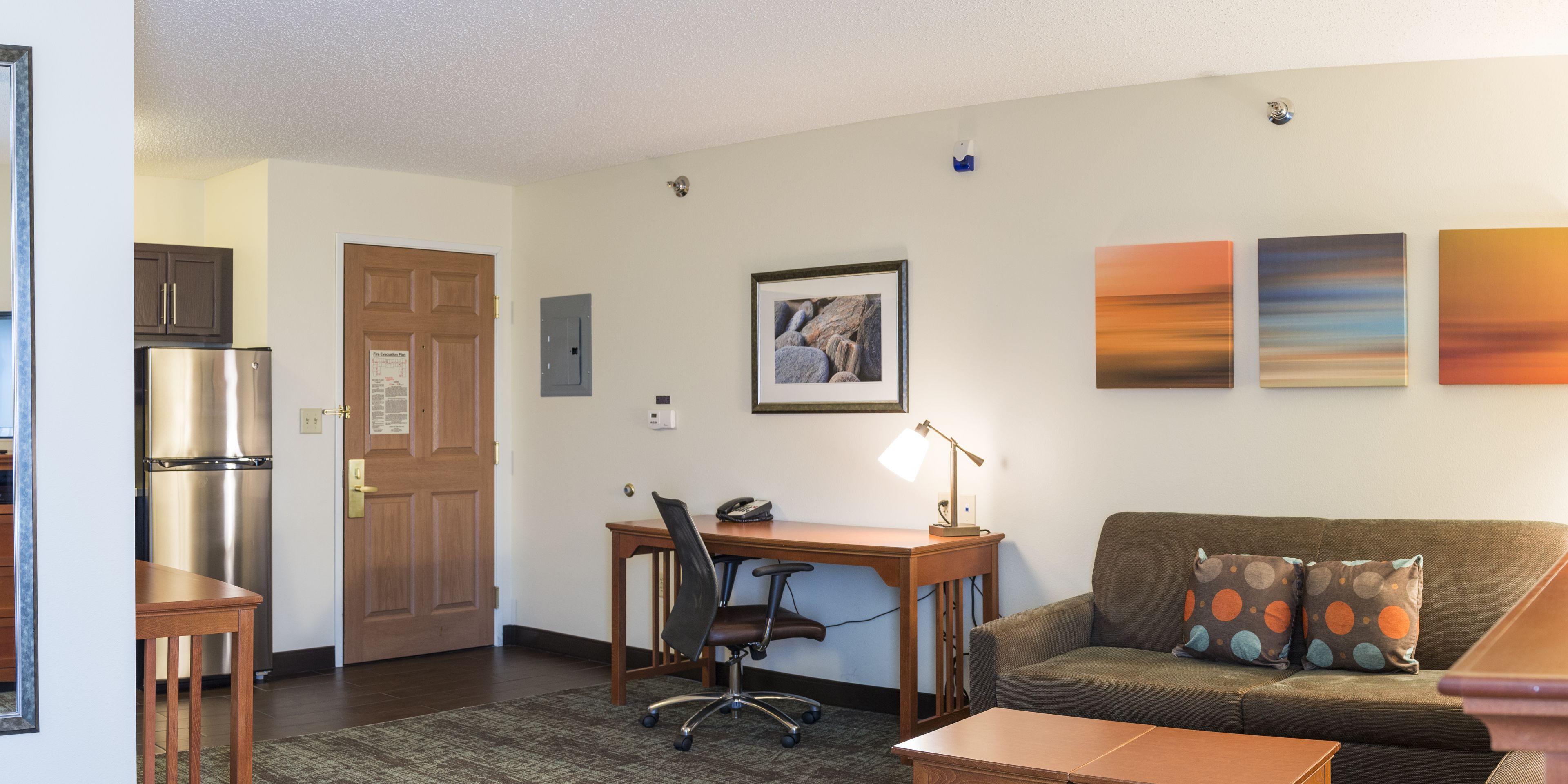 A compact living space with a kitchenette, a wooden desk, a sofa with patterned cushions, and wall art.