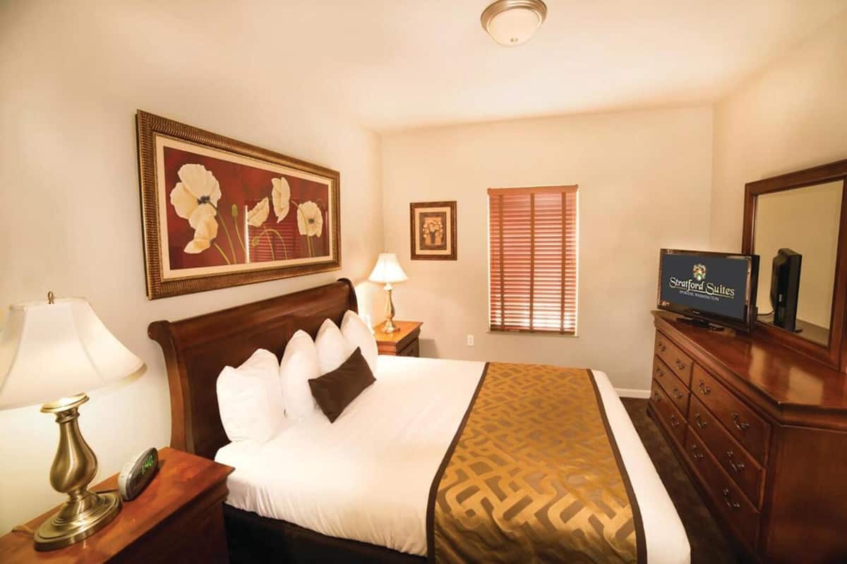A hotel room with a bed, a TV, a dresser, and a painting on the wall.