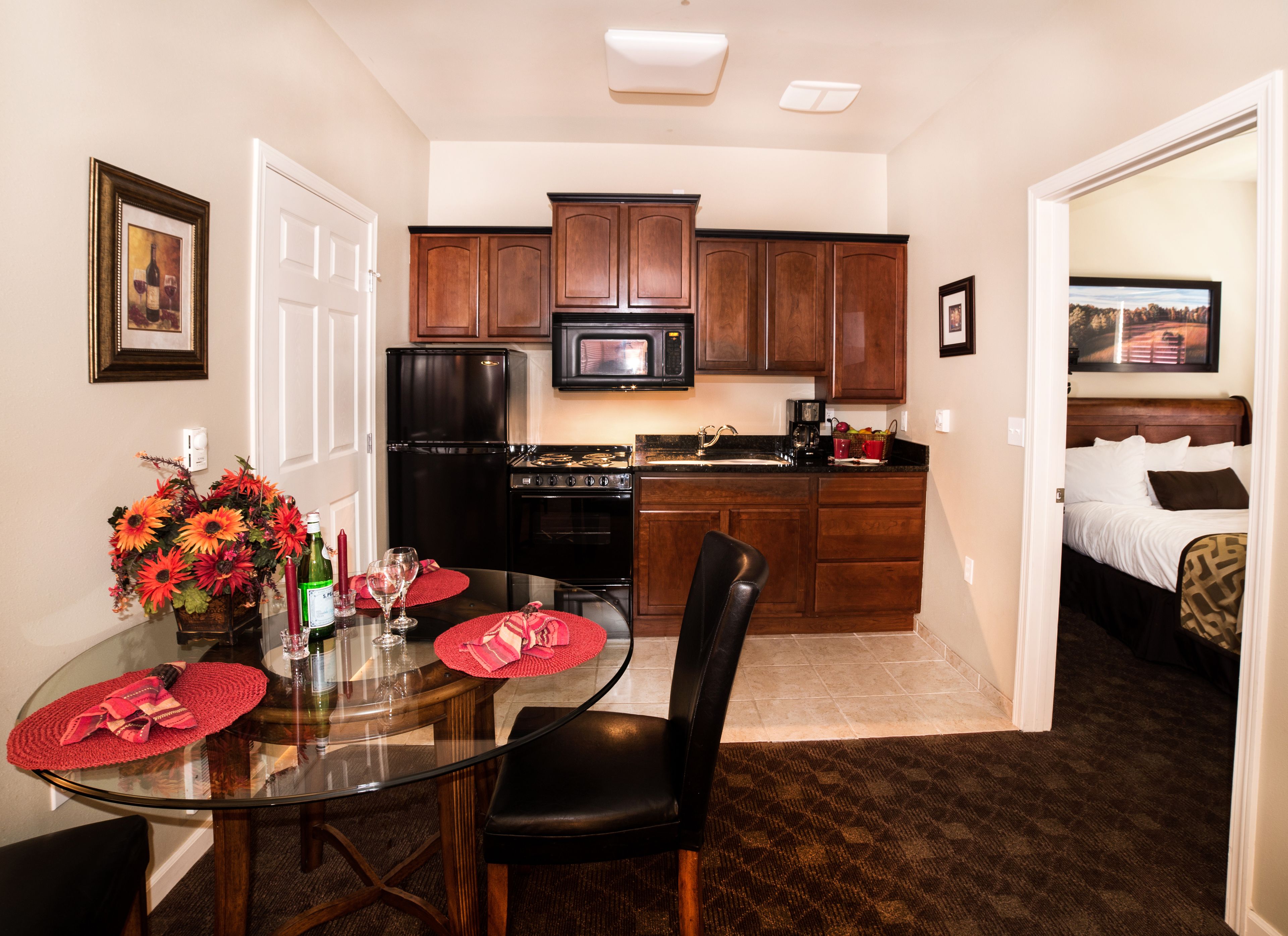 A compact kitchen with a refrigerator, stove, and microwave. There is a dining table with chairs and a vase of flowers. A bed is visible in the adjacent room.