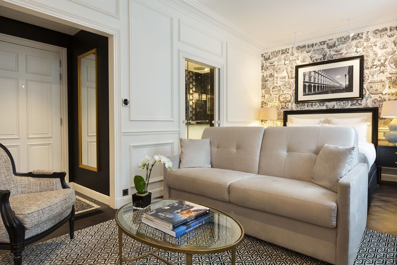 Un salón con un sofá color beige, una mesa de centro redonda de vidrio con libros, una alfombra estampada, un cuadro en la pared y una puerta con espejo.