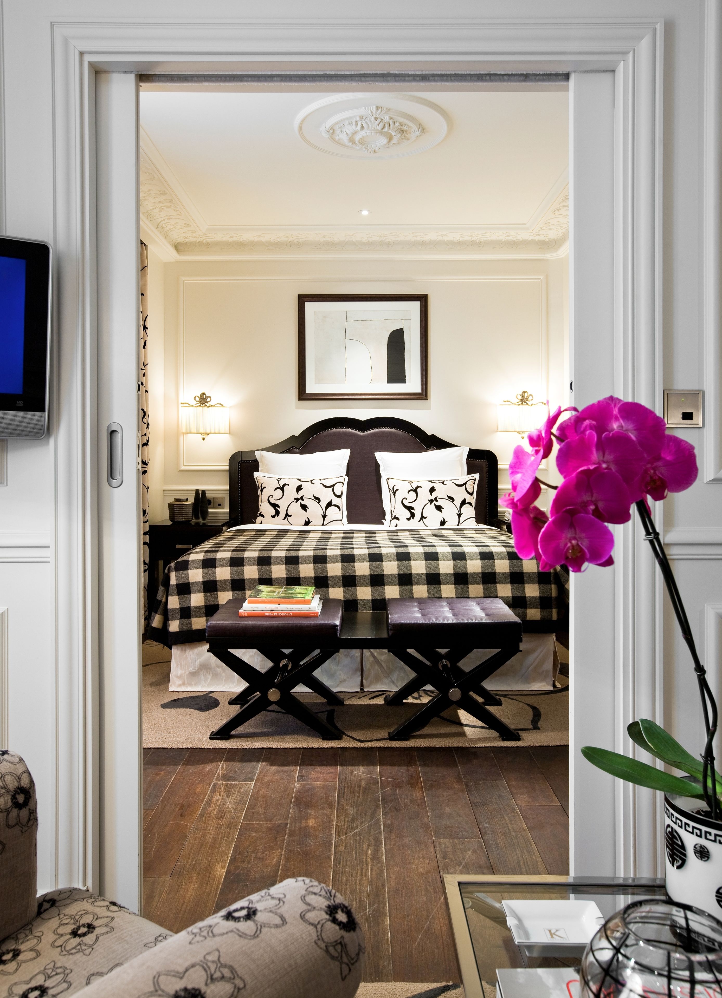 A hotel room with a checkered bedspread, a bedside table with books, a large window, and a view of the room's interior.