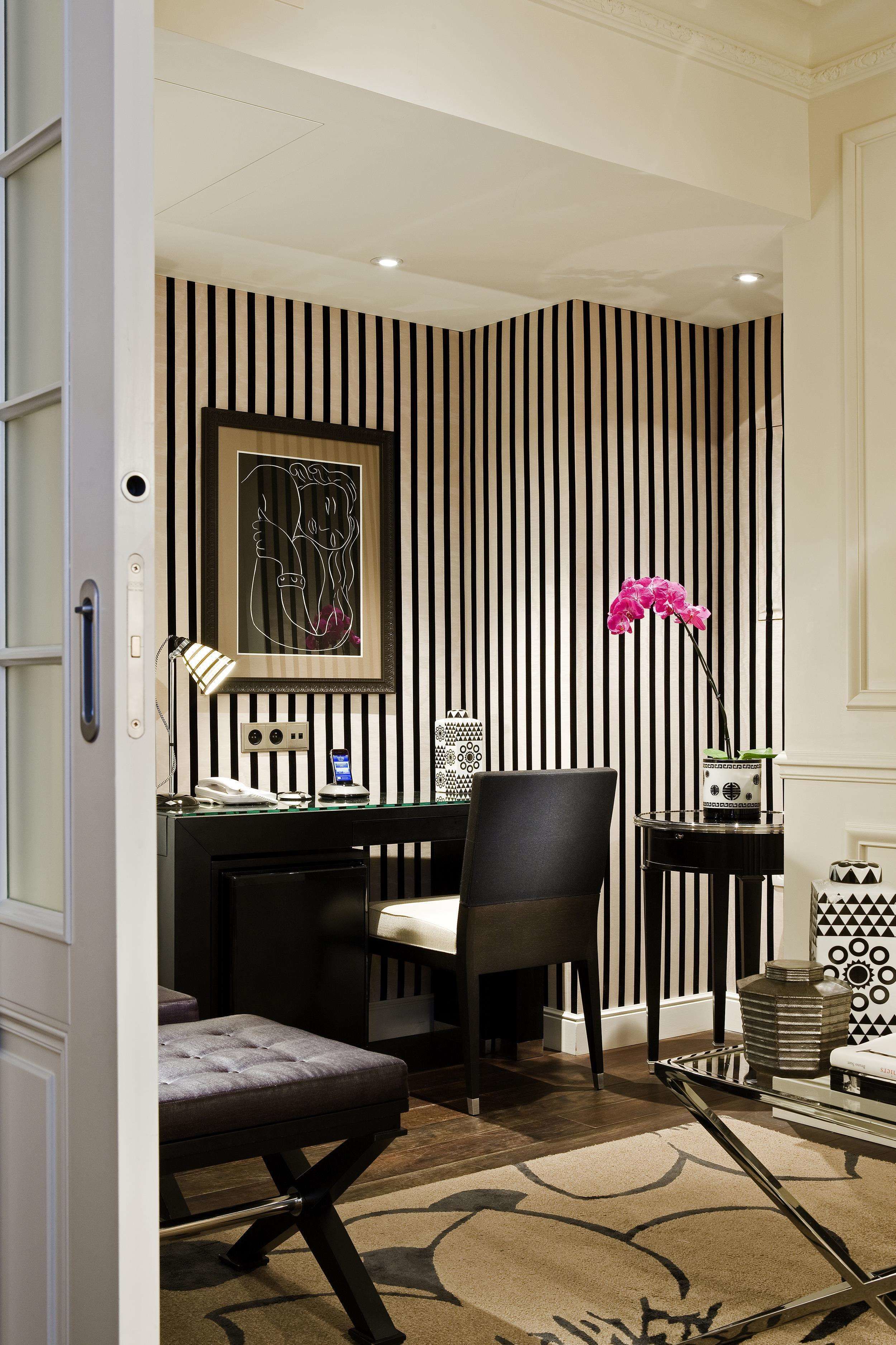 A room with a striped wall, a desk with a chair, a telephone, a lamp, and a pink orchid.