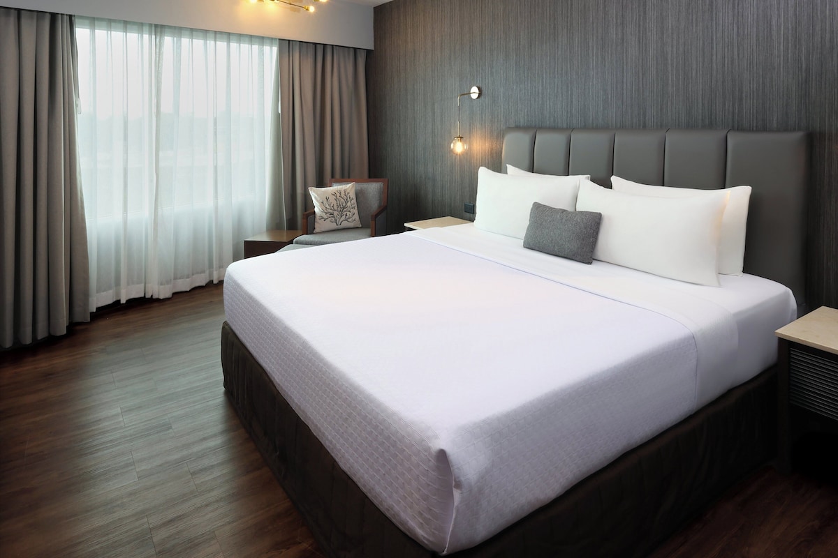A hotel room with a large bed, a nightstand, a chair, and a wall-mounted lamp.
