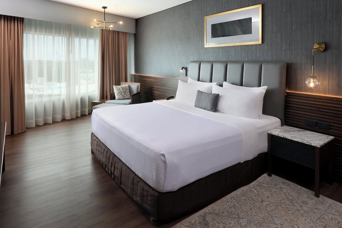 A modern hotel room with a large bed, a nightstand, a wall-mounted lamp, and a view of the city through sheer curtains.