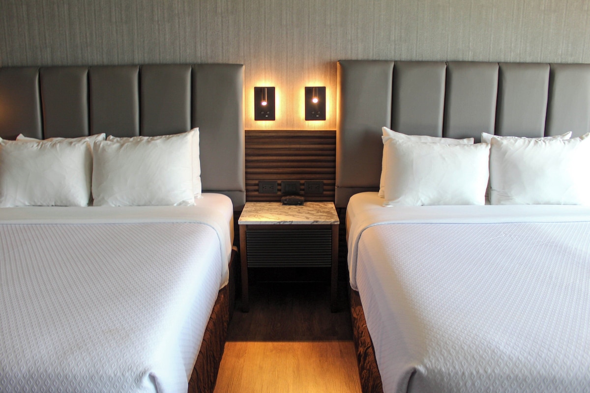 A hotel room with two beds, a wooden nightstand, and a wall-mounted light fixture.