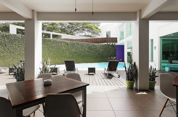 Een terras buiten met een houten tafel en stoelen, een zwembad en potplanten.