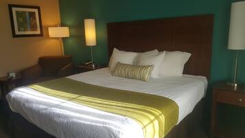 Una habitación de hotel con una cama grande, cabecera, mesita de noche con lámpara, una silla e imagen enmarcada en la pared.
