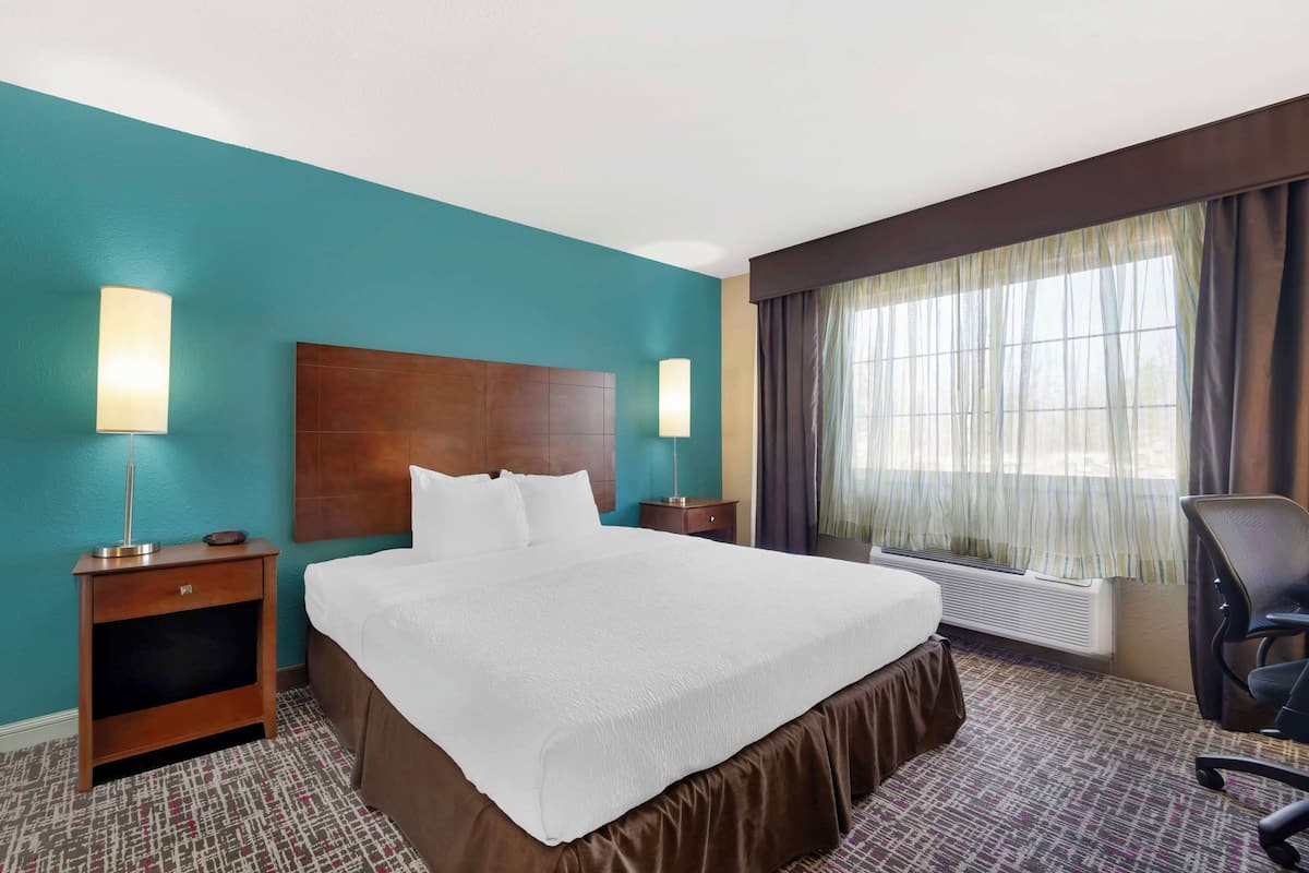 A hotel room with a large bed, two bedside tables, a desk with a chair, a window with curtains, and a wall heater.