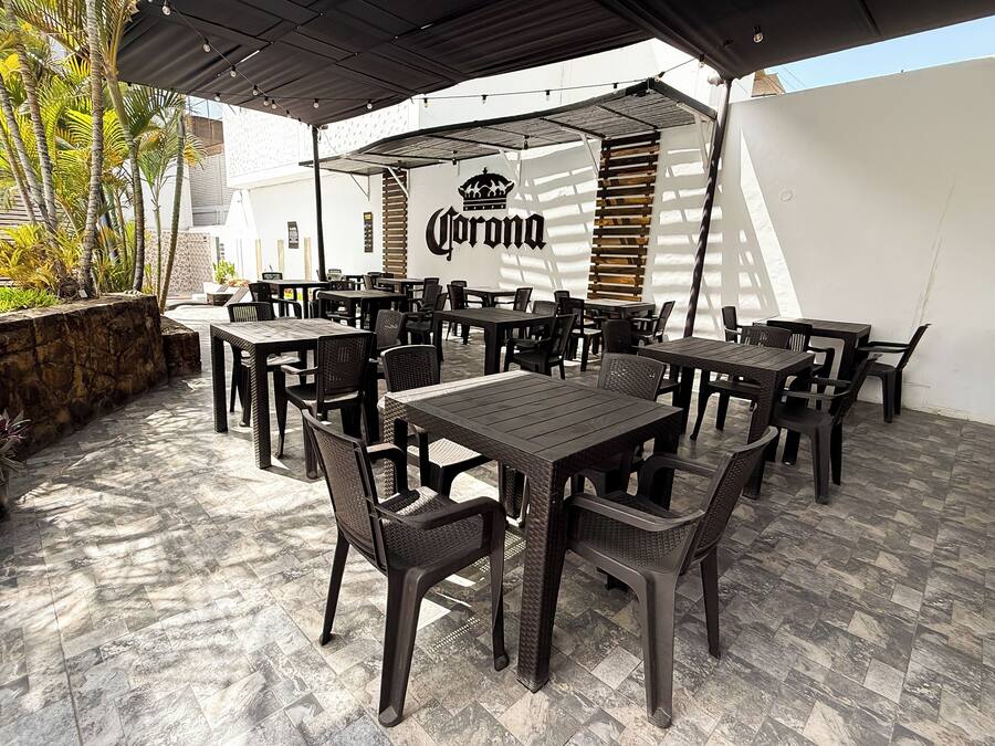 En utomhusplats med flera bord och stolar, en Corona-logotyp på väggen och en stenmur med växter.