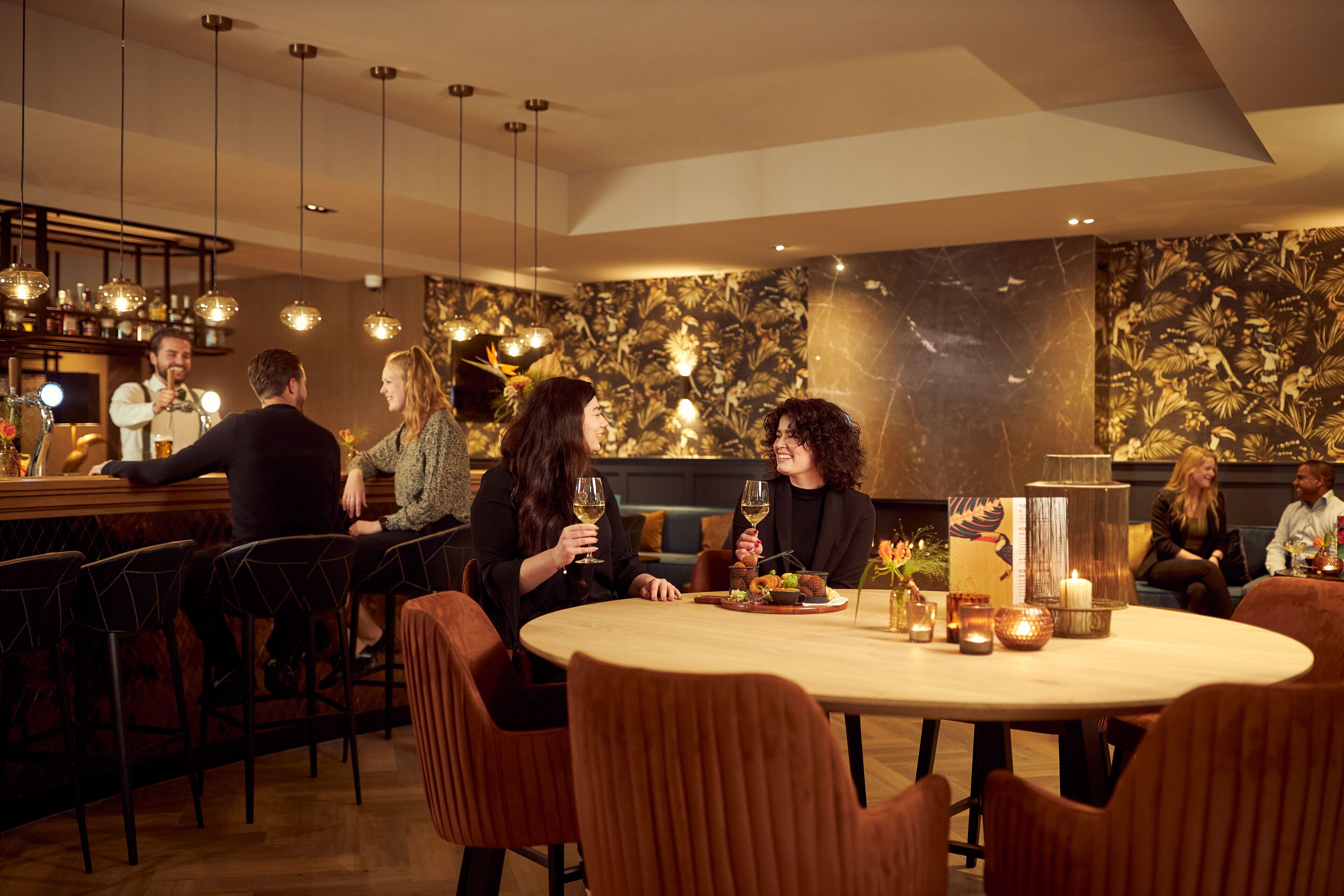 A restaurant with a bar area, round tables, and modern lighting fixtures.