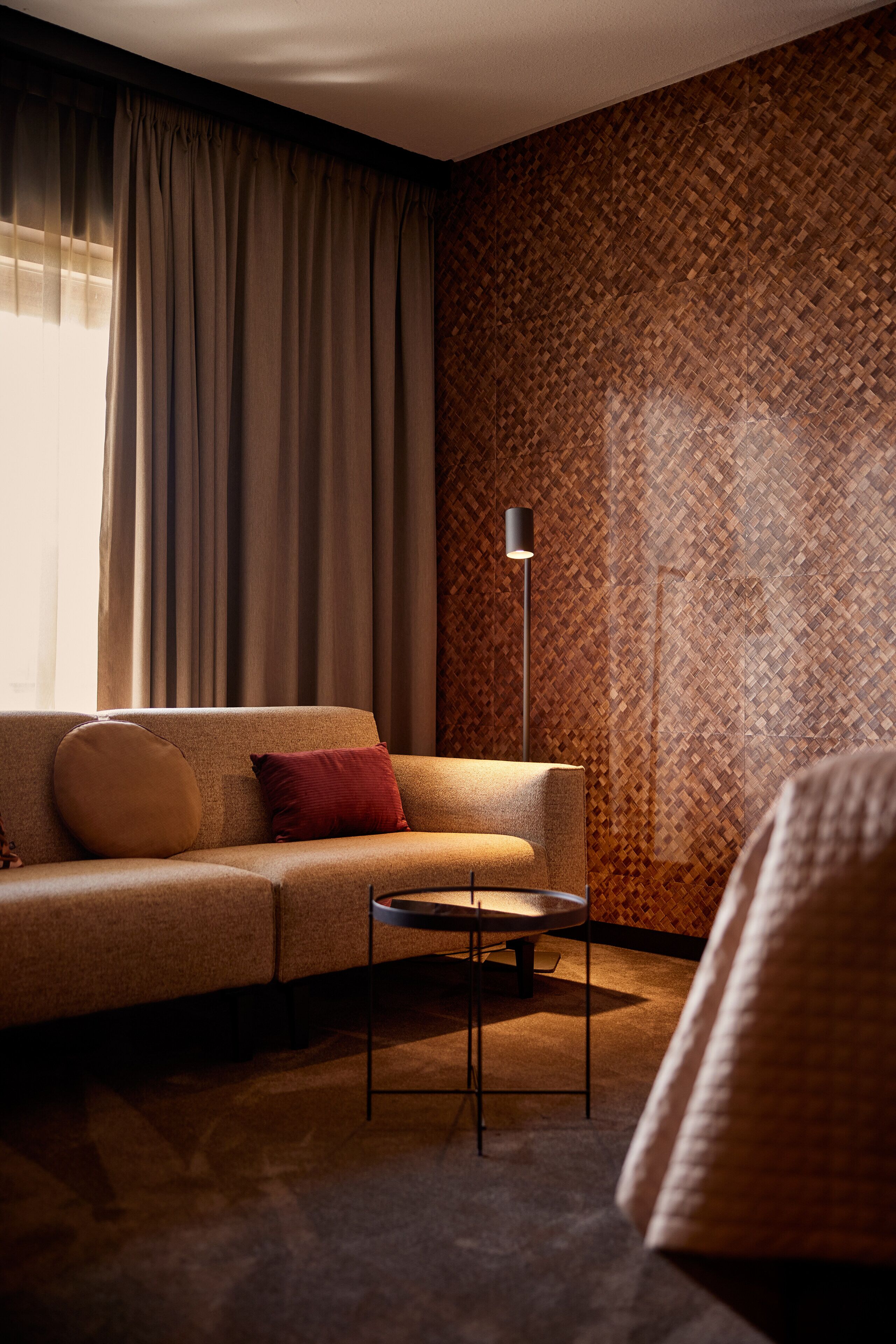 A modern living room with a sofa, a small table, and a floor lamp. The room has a patterned wallpaper and a large window with curtains.