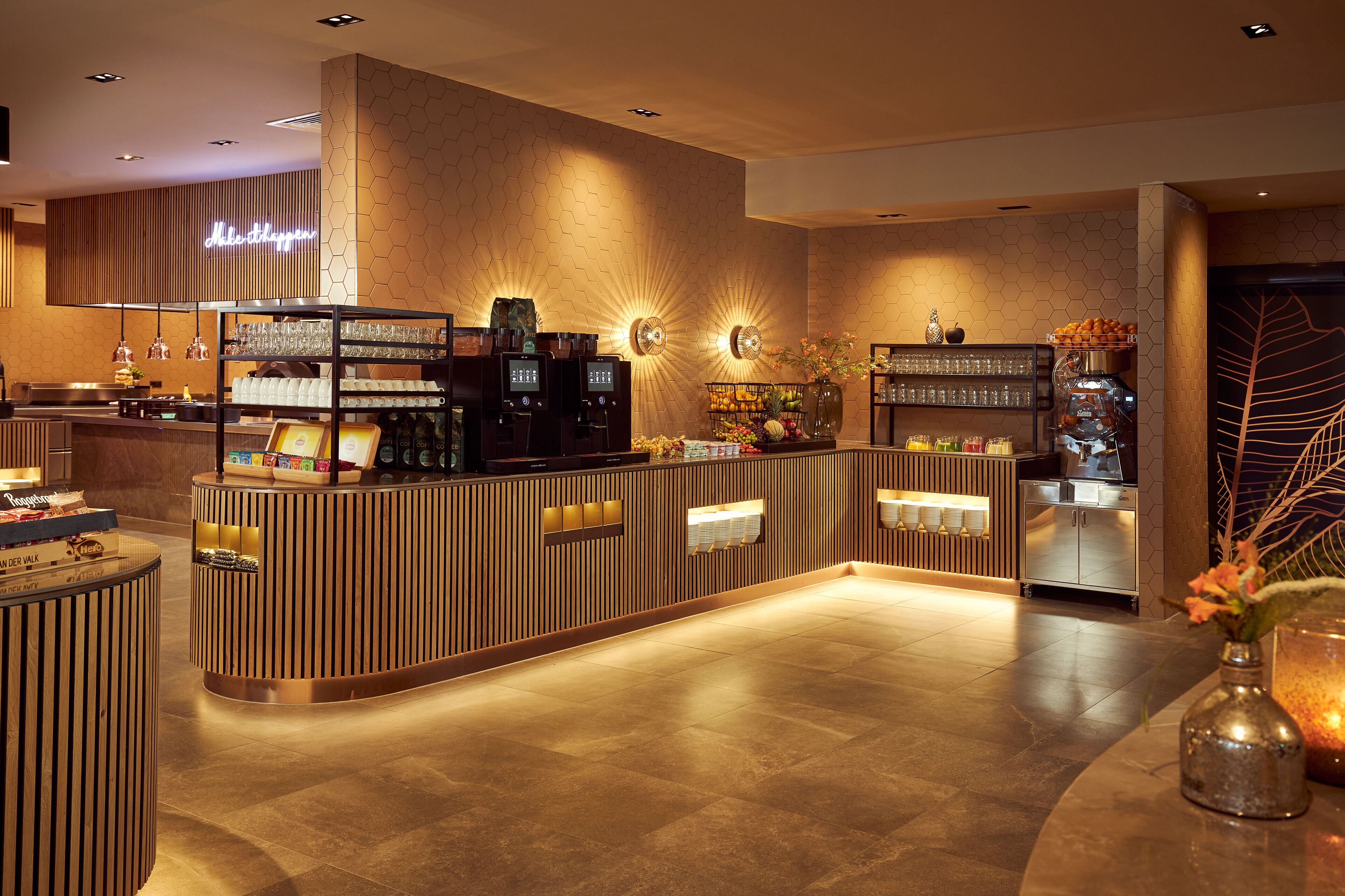 A modern hotel lobby with a reception desk, coffee machines, and a wall-mounted sign.