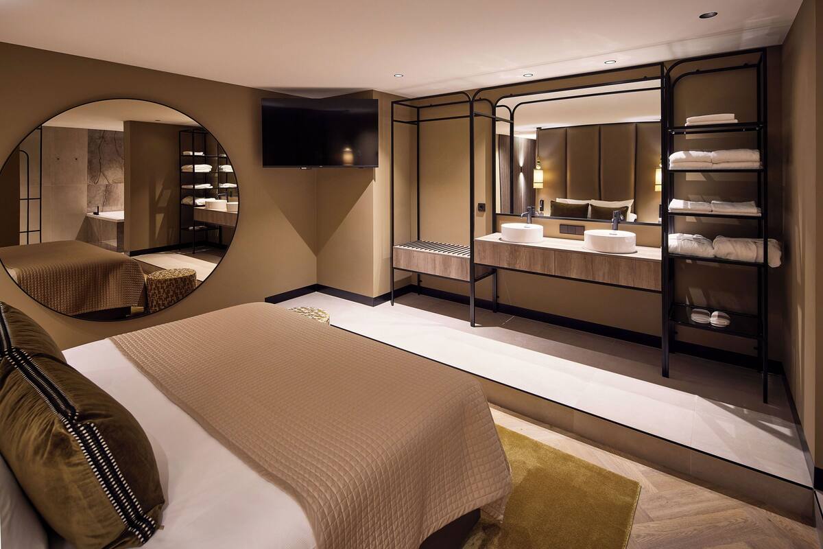 A modern hotel room with a large bed, a round mirror, a vanity with a sink, and a walk-in closet with shelves.