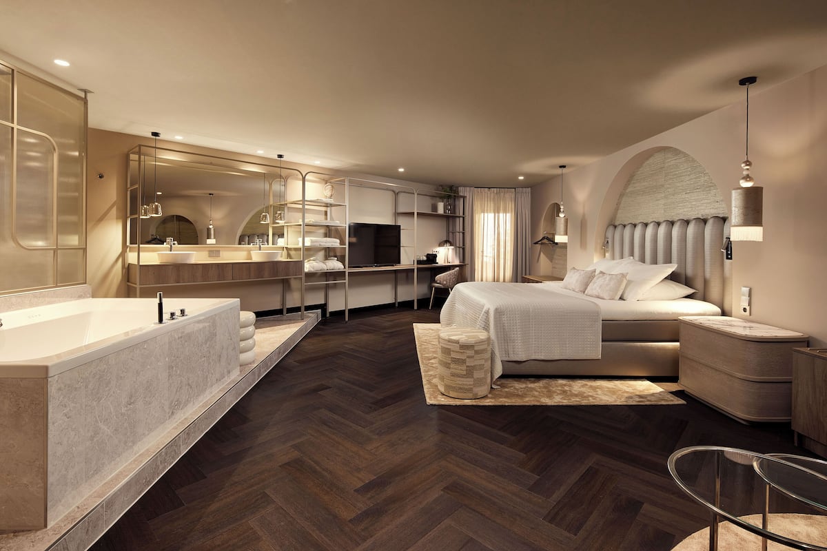 A modern hotel room with a large bed, a separate bathroom with a bathtub, a desk with a computer, and a large mirror.