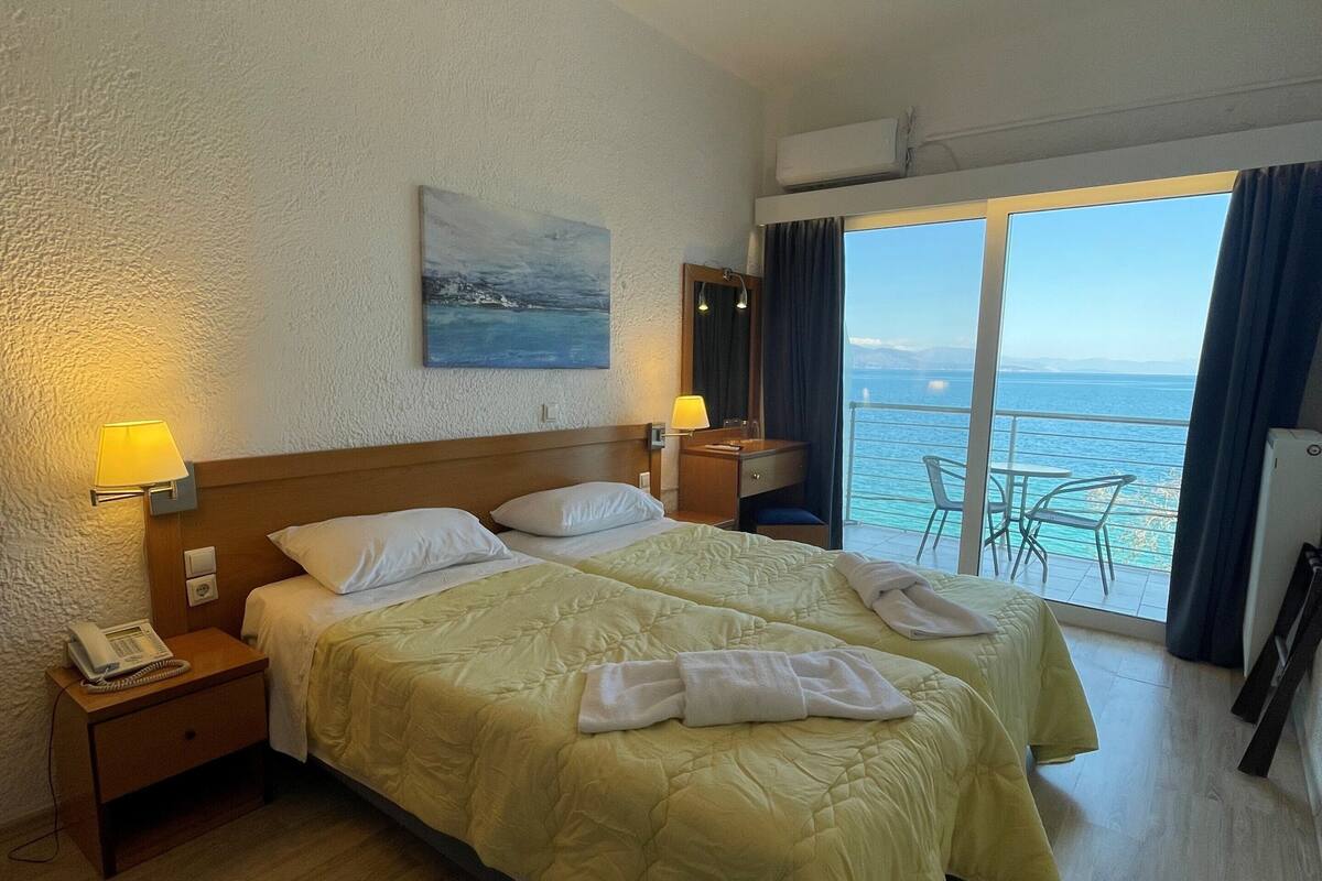 A hotel room with a large bed, two bedside lamps, a painting on the wall, and a view of the sea through the window.
