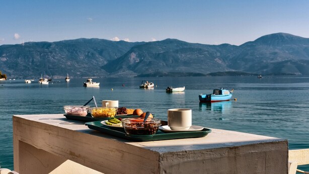 A table with breakfast items and a view of boats on a lake with mountains in the background. - Kanelli Beach (Aigialeia)