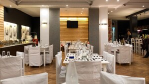 A restaurant with tables set for dining, white chairs, and a wooden wall with artwork. - Kanelli Beach (Aigialeia)