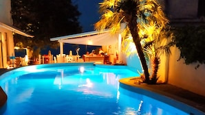 A swimming pool with a blue hue, surrounded by outdoor seating and a palm tree. - Kanelli Beach (Aigialeia)