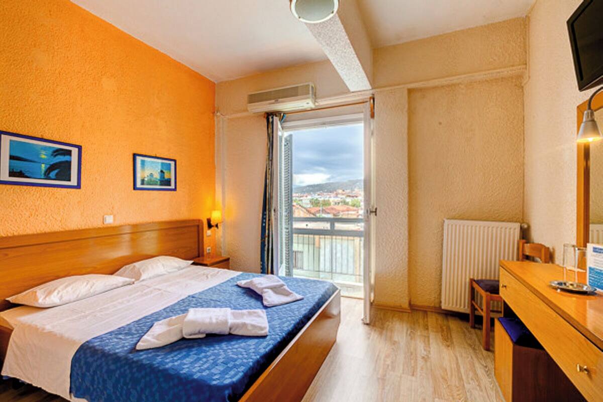 A hotel room with a bed, a desk, a chair, a window with a view, and a radiator.