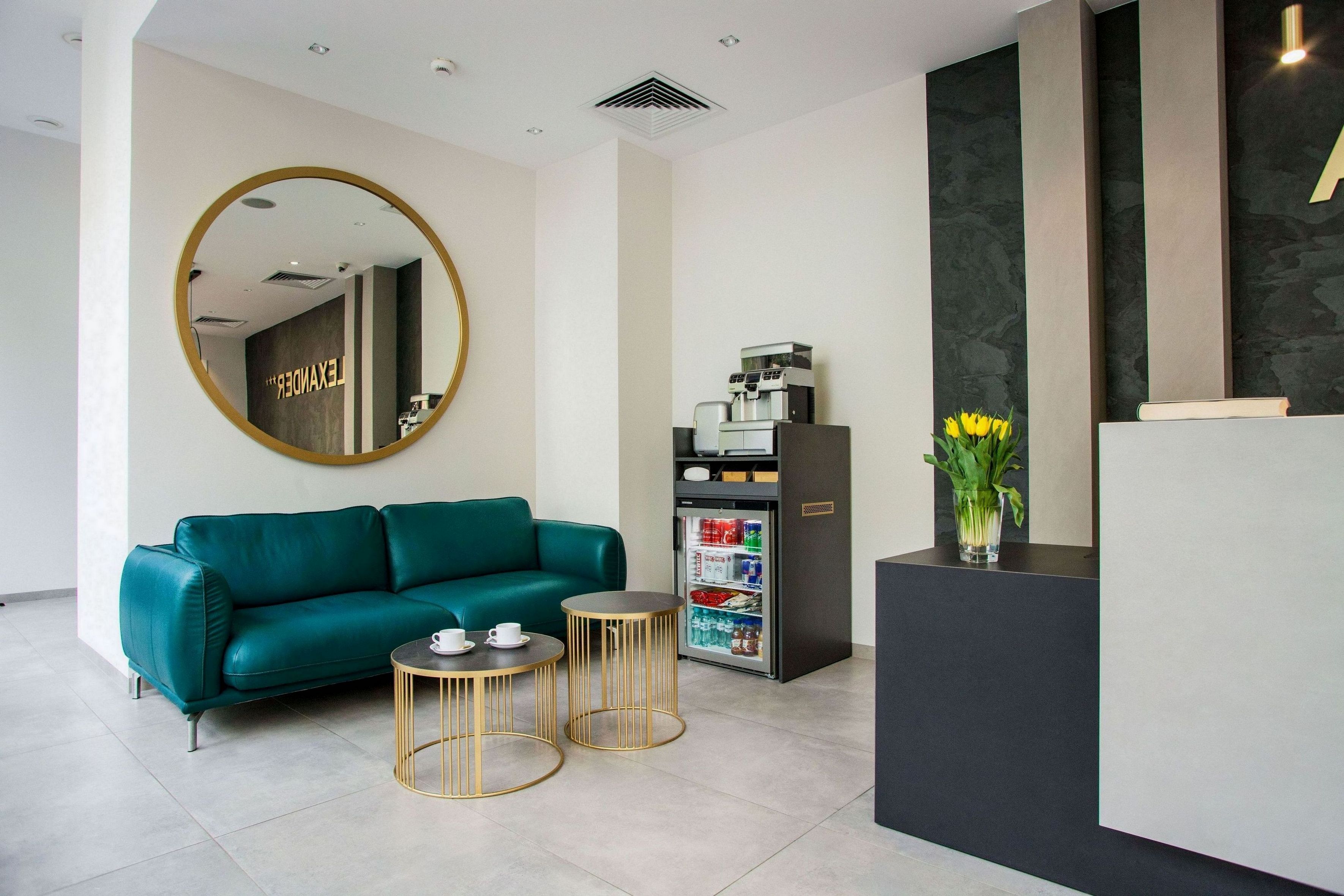 Un área de recepción con un sofá color turquesa, dos mesas con detalles dorados, una máquina de café y un refrigerador.