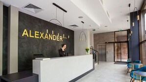 A modern reception desk with a nameplate reading "ALEXANDER" and a receptionist. There are blue chairs and a staircase in the background.